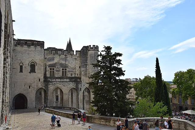 The crenellated stone walls and towers of the Palais des Papes seen from the square, with cypress trees and visitors