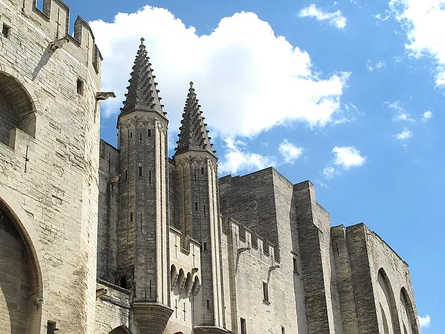 Two pointed stone pinnacles and the crenellated walls of the Palais des Papes against a blue sky with white clouds